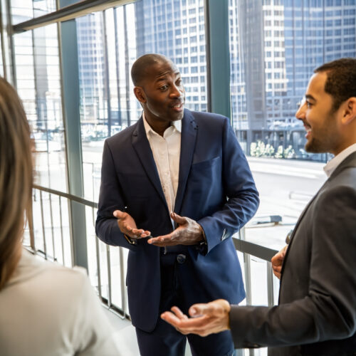 Two executives in animated hallway chat, modern Manhattan office. Huge windows reveal Hudson River. Speaker in blue shirt gestures enthusiastically; listener in white nods, pleased. Blurred background: diverse employees, standing desks, colorful posters. Bright, airy vibe. Shallow focus on foreground figures, capturing innovation and success.