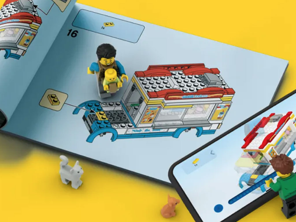 Two Lego block adults, a lego block baby and lego cat and dog looking at a copy of the Lego design book and digital copy of the book on a phone.